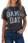 A woman wearing a dark charcoal gray t-shirt with 'GAME DAY' printed in camouflage-patterned letters, paired with blue jeans.